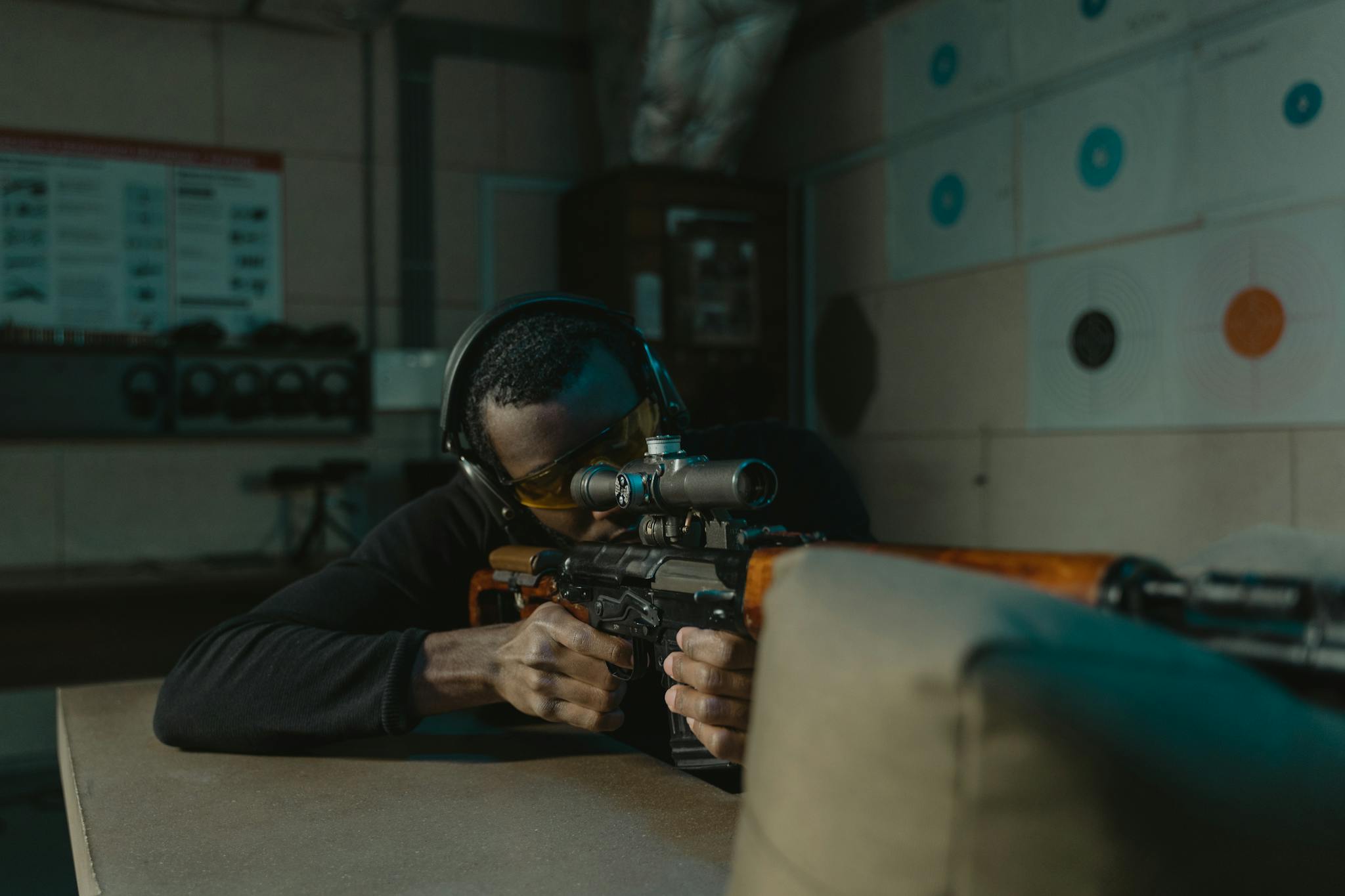 A determined man wearing ear and eye protection aims a rifle at a target in an indoor shooting range.