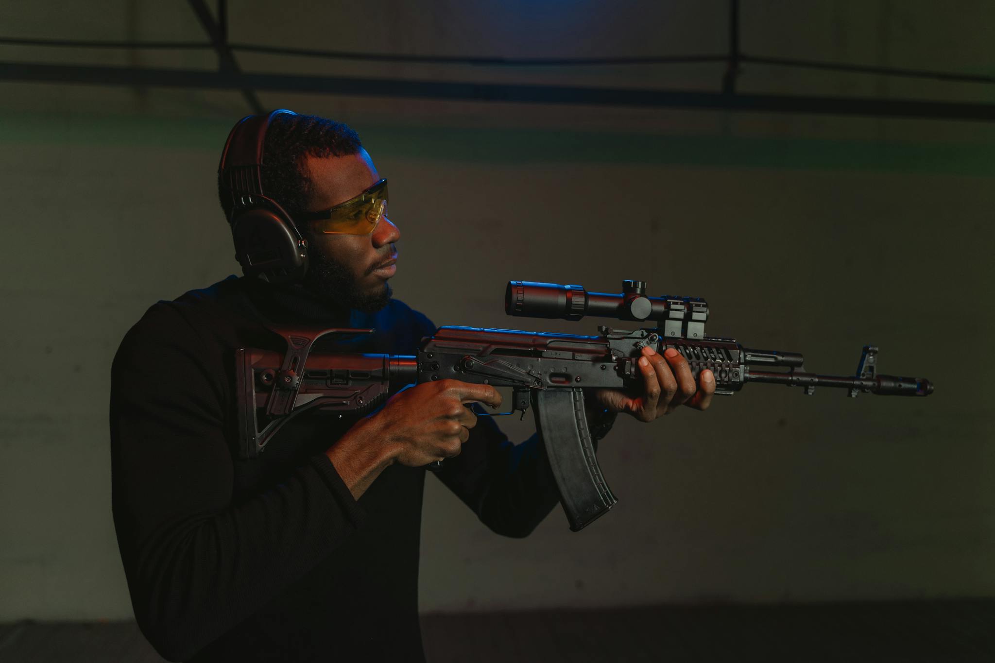 An adult man wearing ear and eye protection aiming a rifle indoors.