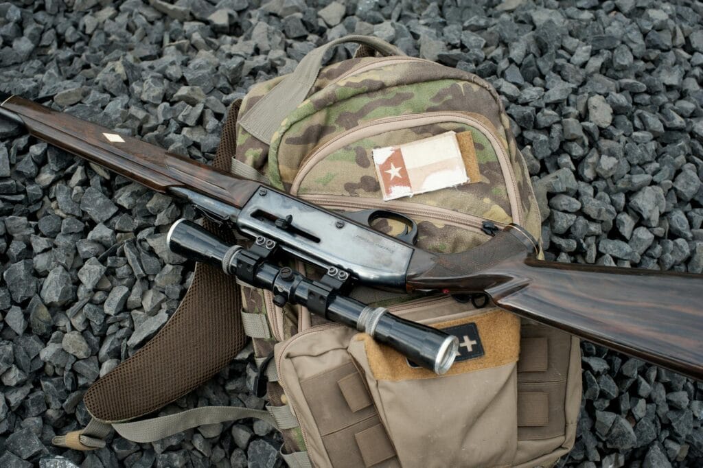 Camouflage backpack with sniper rifle on pebbles, symbolizing military preparedness.