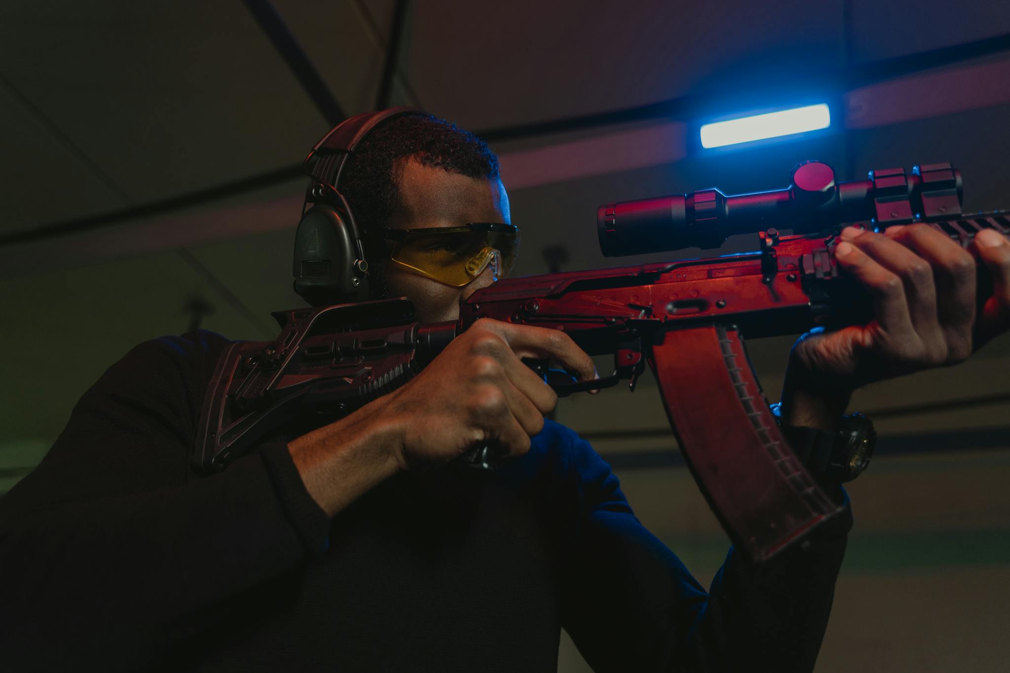 Man in safety glasses holding a sniper rifle at an indoor shooting range. Precise aim and focus displayed.