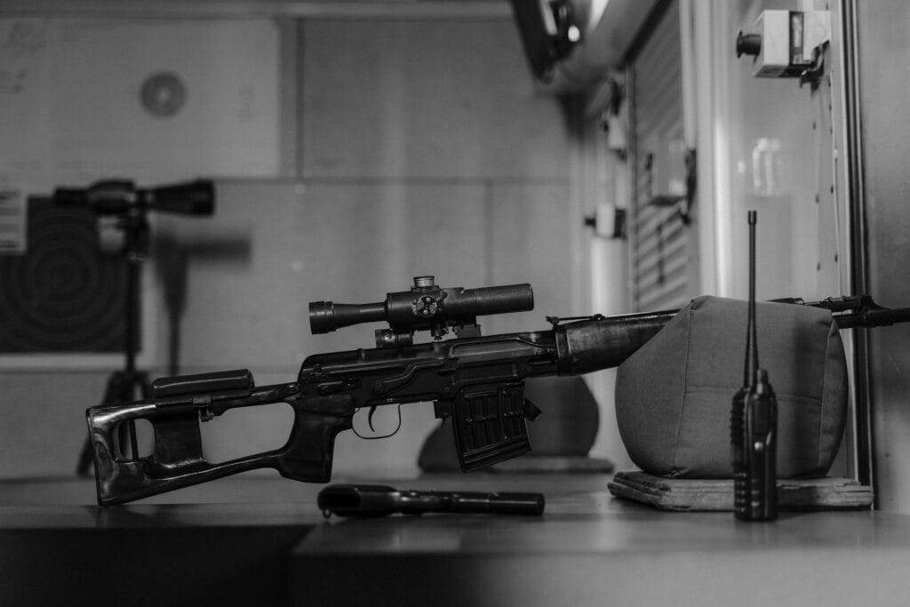 Monochrome image of a sniper rifle on a table indoors with a walkie talkie and scope.